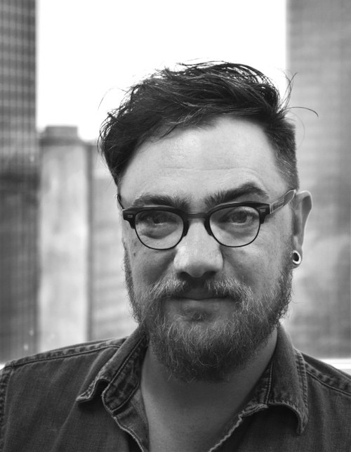 Black-and-white photograph of Benjamin Painter, bearded and wearing round glasses, against city buildings